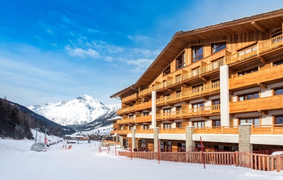 View of the exterior of the Hotel Saint Charles ****, Val Cenis, France, next to snow-covered nursery slopes with panoramic mountain scenery and blue skies above