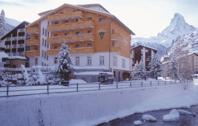 View of the wood-clad exterior of the 3 star superior Hotel Perren in Zermatt, Switzerland, in winter, looking across a river, with snow on the ground and trees, and view of the Matterhorn in the distance
