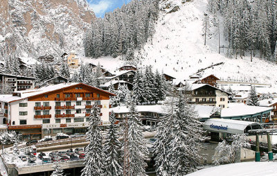 View of the chalet-style exterior of the 4 star Hotel Aaritz and the village of Selva, Italy, in winter, with snow-covered mountain scenery beyond