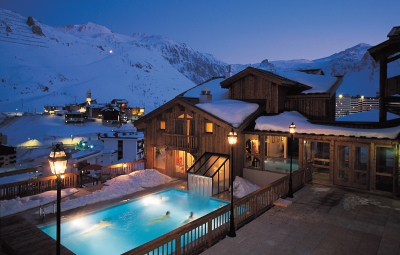 Night-time view of the exterior of the 4 star Hotel Village Montana in Tignes, France, and its heated outdoor swimming pool with snow-covered mountains behind