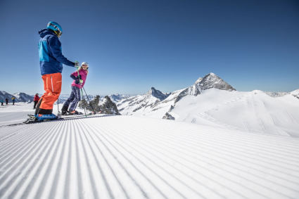 Skiers on a freshly groomed piste on the Hintertux glacier in Austria, with snow-covered Alpine peaks in the background